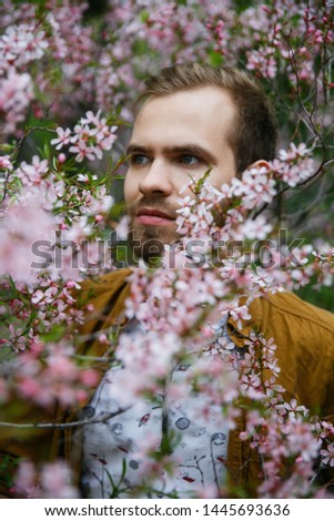Young cute stylish guy dreams in green and lush pink almond flowers on summer morning
