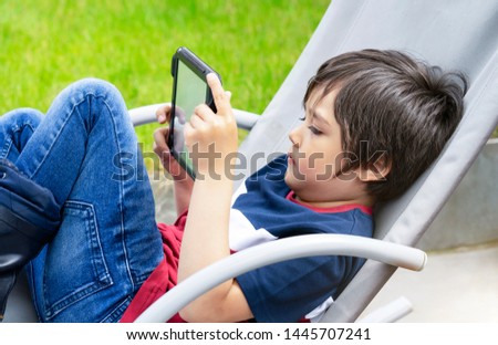 Portrait kid sitting on rocking chair playing game on tablet in the garden,  Child boy having fun watching cartoons on digital taplet,Kid with smiling face playing games on touch pad.