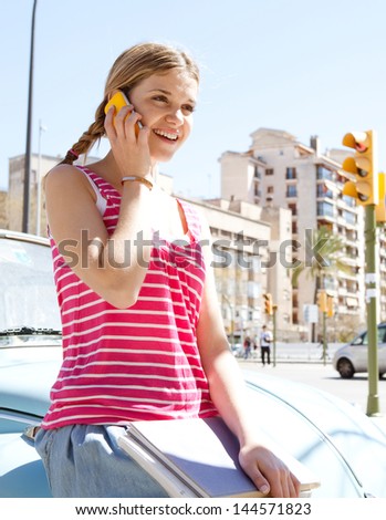 Portrait of a teenager girl having a phone call conversation with her smartphone while sitting on a classic colorful car in a city during a sunny day with a blue sky, smiling.