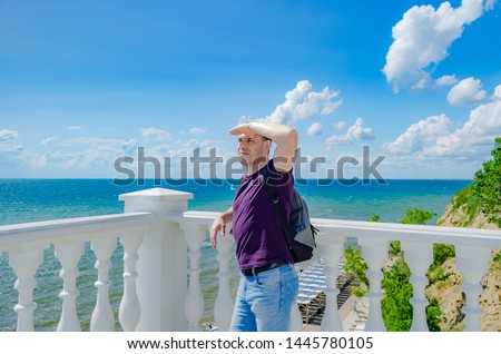 A healthy elderly man of athletic build stands near a white fence with balusters and looks at the blue sea.