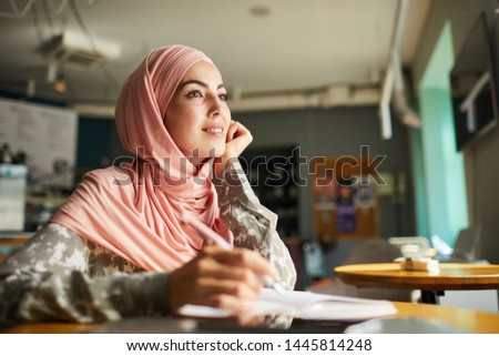 Pretty positive young woman in hijab writing her dreams in diary when sitting at table in cafe