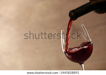 Pouring red wine into glass from bottle against blurred beige background, closeup. Space for text