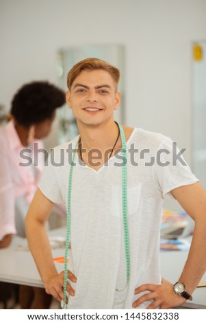 Cheerful mood. Happy smiling blond young man with a measuring tape on the shoulders standing in a fashion designers studio