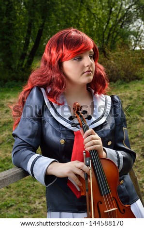 A teenage girl is standing with a violin outdoors. She is wearing an anime costume and a wig.