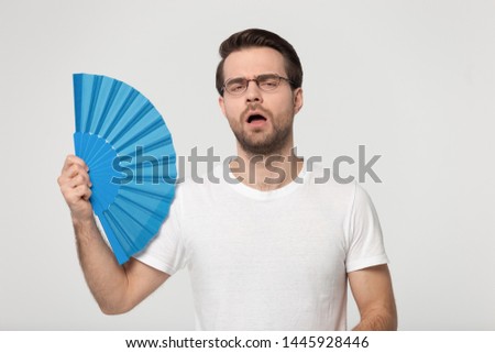 Guy wearing gasses white t-shirt feels overheated holds blue waver fan induces airflow cooling himself reducing heat pose isolated on gray studio background, summer weather, no air conditioner concept Royalty-Free Stock Photo #1445928446