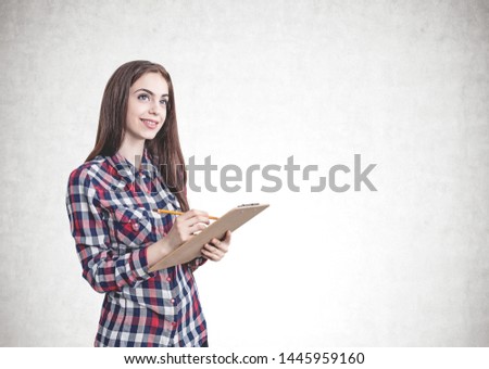 Portrait of smiling young woman with long brown hair wearing casual clothes and writing in clipboard standing near concrete wall. Mock up