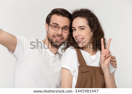 View from webcam girl smiling showing victory sign hand gesture makes photo with handsome boyfriend in glasses who hugs beloved girlfriend, shooting together look at camera having fun using smartphone