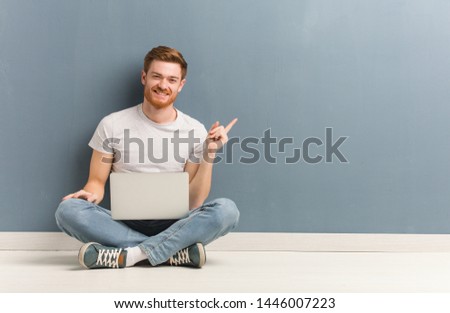 Young redhead student man sitting on the floor pointing to the side with finger. He is holding a laptop.