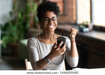 Excited african american millennial tattoo girl in glasses hold smartphone feel euphoric reading good news, overjoyed happy black biracial young woman use cellphone triumph win lottery online Royalty-Free Stock Photo #1446009680