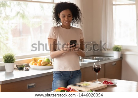 African woman standing in kitchen distracted from cooking preparing dinner received sms read message uses phone, searching recipe, taking picture of vegetable dish social networks user blogger concept