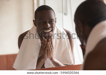 Black man towel on shoulders standing in bathroom look in mirror washes face touch skin after shaving or applying cream aftershave lotion feels satisfied, morning routine, personal hygiene, skincare Royalty-Free Stock Photo #1446020366