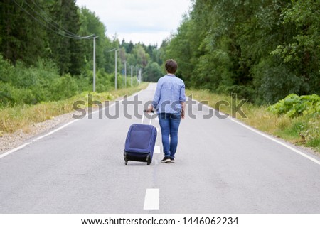 Middle-aged Woman tourist with a travel bag on wheels walks along the narrow rural road into the distance. Travel concept