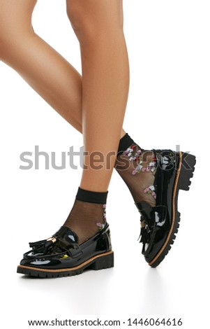 Cropped side shot of lady's legs in black nylon socks, adorned with versicolored floral insert. The girl is wearing patent leather shoes with ribbed soles and tassels, posing on white background.