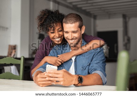 Smiling young couple embracing while looking at smartphone. Multiethnic couple sharing social media on smart phone. Smiling african girl embracing from behind her happy boyfriend while using cellphone Royalty-Free Stock Photo #1446065777