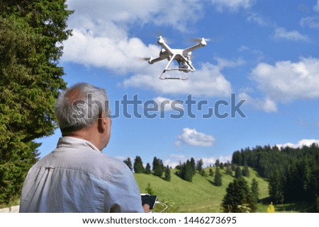Senior man with white drone. Man navigates unmanned aerial vehicle among nature. Retired person leisure activity. Pine forest, green meadows and blue sky at background