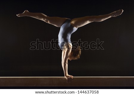Side view of a female gymnast doing split handstand on balance beam against black background