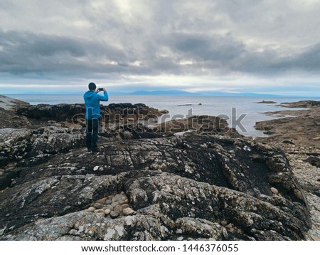 Tourist in a blue jacket taking picture on his smart phone of dramatic landscape, Galway bay, Burren in the background.
