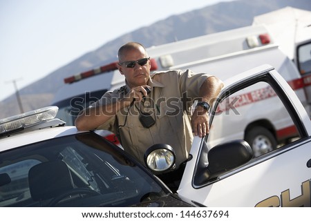 Police officer using two way radio by police car with ambulance in background