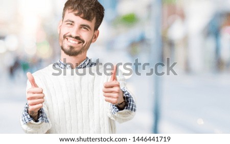 Young handsome man wearing winter sweater over isolated background approving doing positive gesture with hand, thumbs up smiling and happy for success. Looking at the camera, winner gesture.
