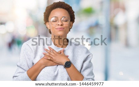 Young beautiful african american woman wearing glasses over isolated background smiling with hands on chest with closed eyes and grateful gesture on face. Health concept.
