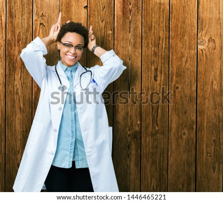 Young african american doctor woman wearing medical coat over isolated background Posing funny and crazy with fingers on head as bunny ears, smiling cheerful