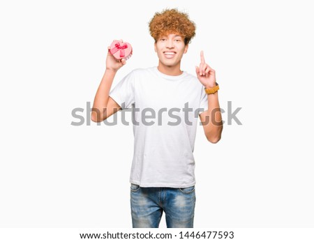 Young handsome man holding heart box as gift of valentine's day surprised with an idea or question pointing finger with happy face, number one