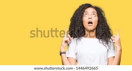 Young beautiful girl with curly hair wearing casual white t-shirt amazed and surprised looking up and pointing with fingers and raised arms.