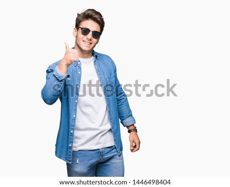 Young handsome man wearing sunglasses over isolated background doing happy thumbs up gesture with hand. Approving expression looking at the camera showing success.