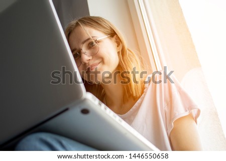 young pretty girl sitting near the window with a laptop, she uses the computer at home on the windowsill, a woman freelance, copy space for text
