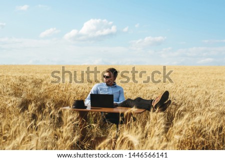 Businessman sitting holding sunglasses at work table with a laptop on her in the wheat field
