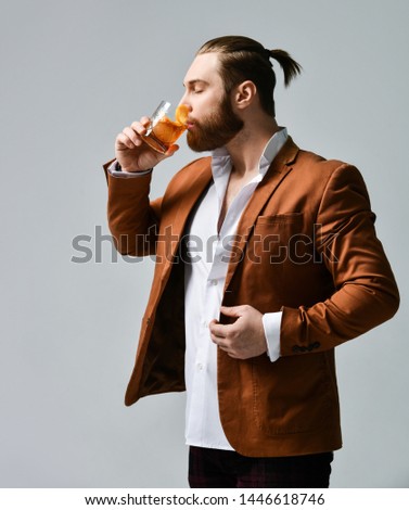 Young stylish modern man bearded barfly in fashion suit tries takes a sip of long awaited delicious cocktail drink with orange on light gray