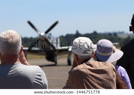 People in a crowd watch an airshow.  Vintage plane in soft focus as a crowd photographs and watches an airshow in the summer.  Happy People gather as an airshow is enjoyed.