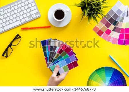 Designer office desk with tools, pallet in hands, coffee, keyboard and glasses on yellow background top view
