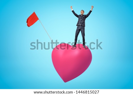 Young happy businessman raising arms up on big pink heart with red flag on blue background. Banking and finance. People and objects. Feelings and emotions.