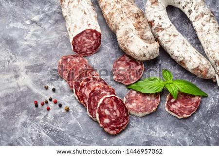 A lot of pieces of dried sausage with pepper spice and basil knife on a dark stone background. Minimalistic composition of food.