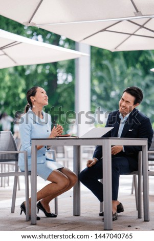 A pair of young Asian business people (a man and a woman) have a business discussion together at a meeting. They are both professionally dressed in suits and are speaking animatedly. 