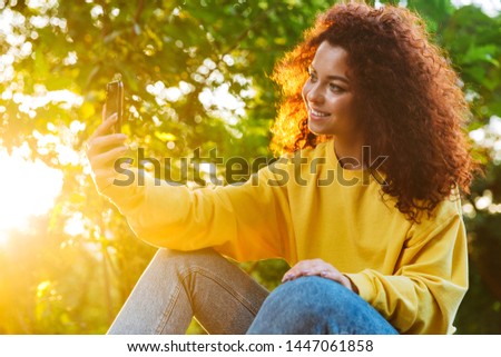 Image of joyful young woman with curly brown hair smiling and taking selfie photo on smartphone while sitting on bench in green park