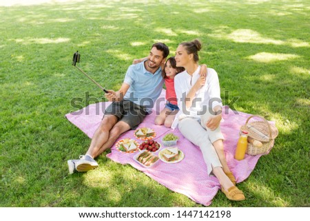 family, leisure and technology concept - happy mother, father and daughter having picnic and taking picture by smartphone on selfie stick at summer park