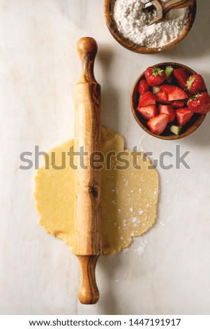 Ingredients for baking summer berry biscuit pie. Bowl of flour, rolled shortbread dough, cutting strawberry and rhubarb over white marble background. Flat lay, space