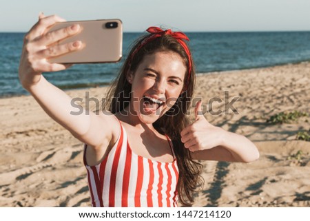 Beautiful cheerful young woman having a good time at the beach on a lovely day, taking a selfie, showing thumbs up