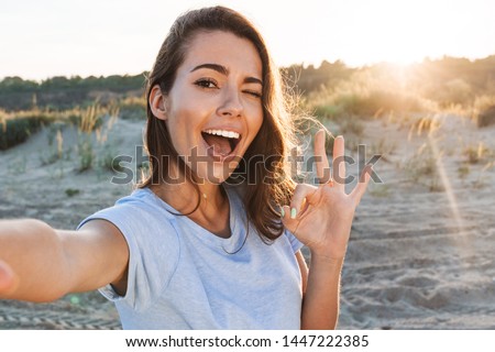 Beautiful cheerful young woman having a good time at the beach on a lovely day, taking a selfie, showing ok gesture