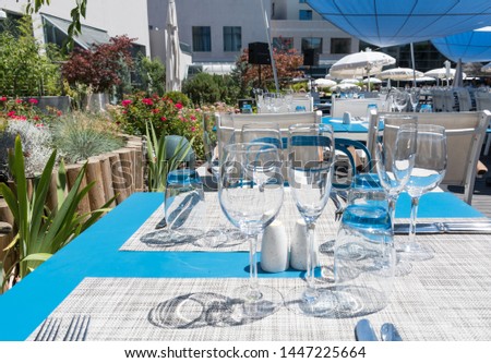 Table setting outdoor terrace. Nobody in the picture just table setup with cutlery and glasses. Pool with sun beds in the background. Blue and wood table, white chairs, summer time and sunshine