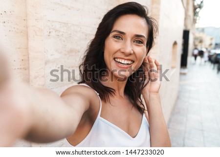 Portrait of happy young woman smiling and taking selfie photo while standing over wall on city street