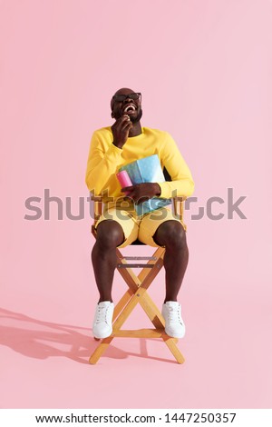 Man watching movie in cinema glasses laughing eating popcorn. Happy black male model in 3d glasses and yellow clothes sitting on director chair watching show on pink background