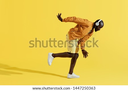 Man in headphones listening music, dancing on yellow background. Full length portrait of handsome black male model in colorful fashion clothes and white earphones in studio