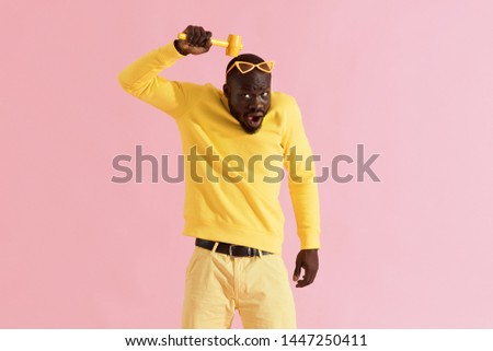 Funny black man beating his head with hammer on yellow background. Colorful portrait of african american male model in yellow fashion clothes behaving silly with mallet in studio 