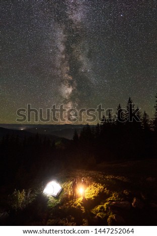 Vertical shot, top view of summer camping in mountains. Young couple hikers standing together beside campfire and tourist tent. On background beautiful night starry sky full of stars and Milky way.