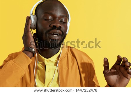 Man in headphones listening music, singing on yellow background. Closeup portrait of black male model in white earphones enjoying to sing to the song