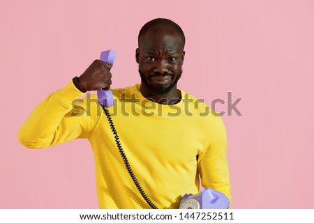 Black man having unpleasant phone call on pink background. Portrait of male with weird "yikes" face expression having uncomfortable talk on purple landline phone in studio Royalty-Free Stock Photo #1447252511