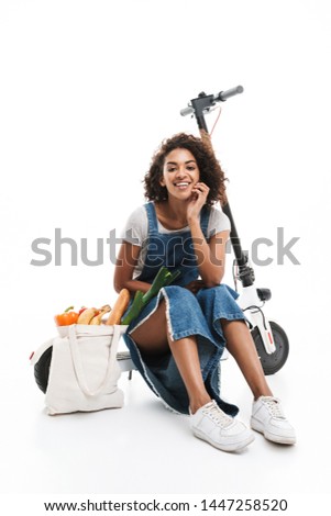 Image of joyful african american woman smiling while sitting on electronic scooter with grocery bag isolated over white background